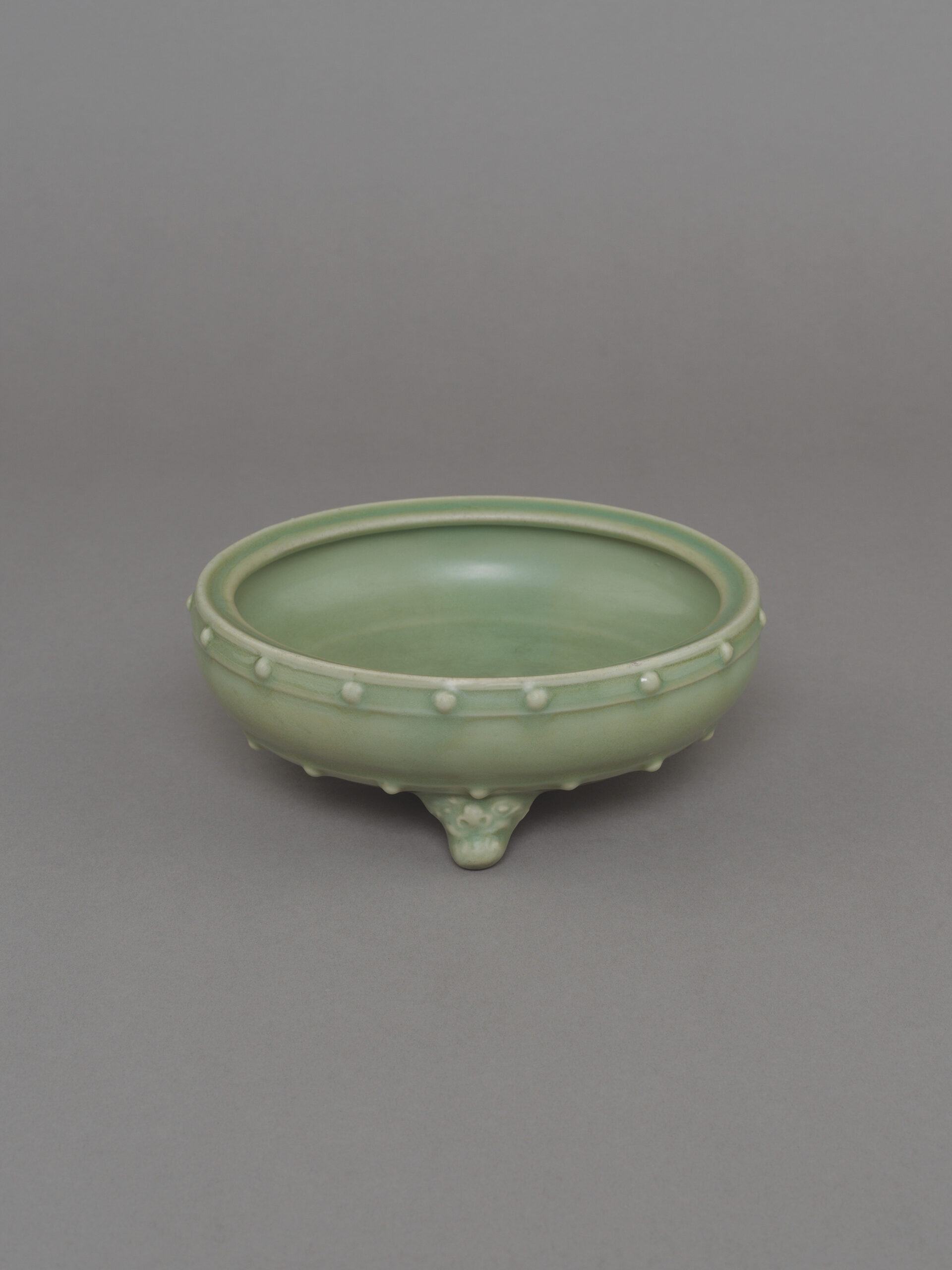 No. 40 - Chinese ceramic Longquan celadon glazed tripod washer, xi, or censer, lu, of circular drum form with two registers of relief drum-nail studs or bosses on three animal-mask feet with gently inverted rim, covered overall in a pale celadon glaze, with knife-cut biscuit foot rim burnt at the edges. 7 1⁄4 inches, 18.5 cm diameter. Southern Song dynasty, Longquan kilns, Zhejiang Province, 12th – 13th century. Japanese wood box, the interior with label inscribed “Sung celadon 3 feet bowl”.